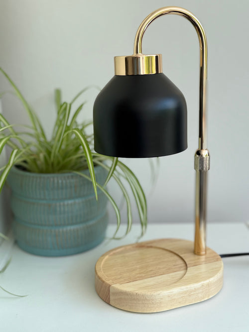 A modern candle warming lamp with a matte black bell-shaped shade, a curved brass-colored metal stem, and a round light wood base, shown on a white surface next to a potted plant.