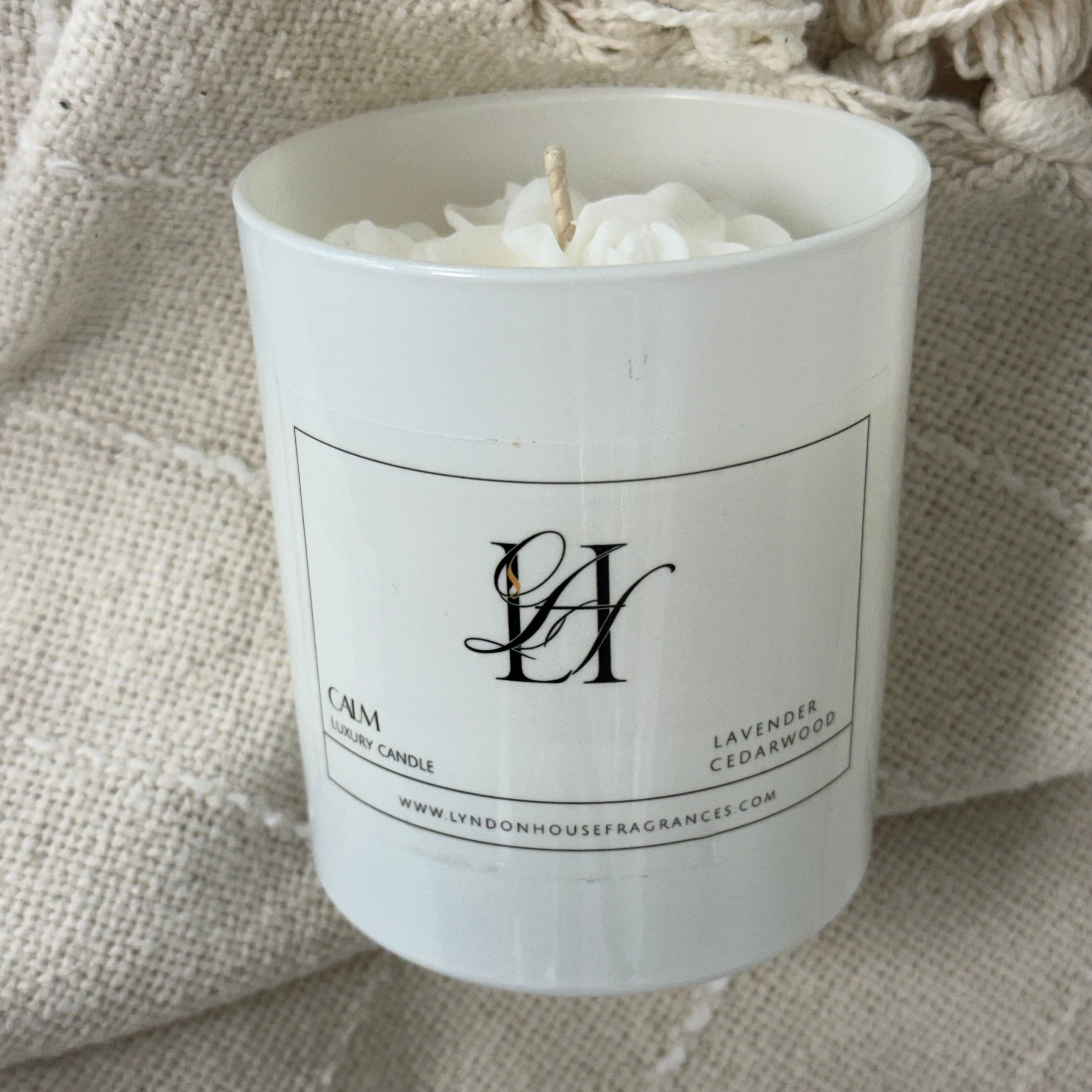 A white glass jar candle with a white wax fill and a single wick, featuring a black printed label with a monogram logo and text. The candle is styled on a textured beige woven blanket.