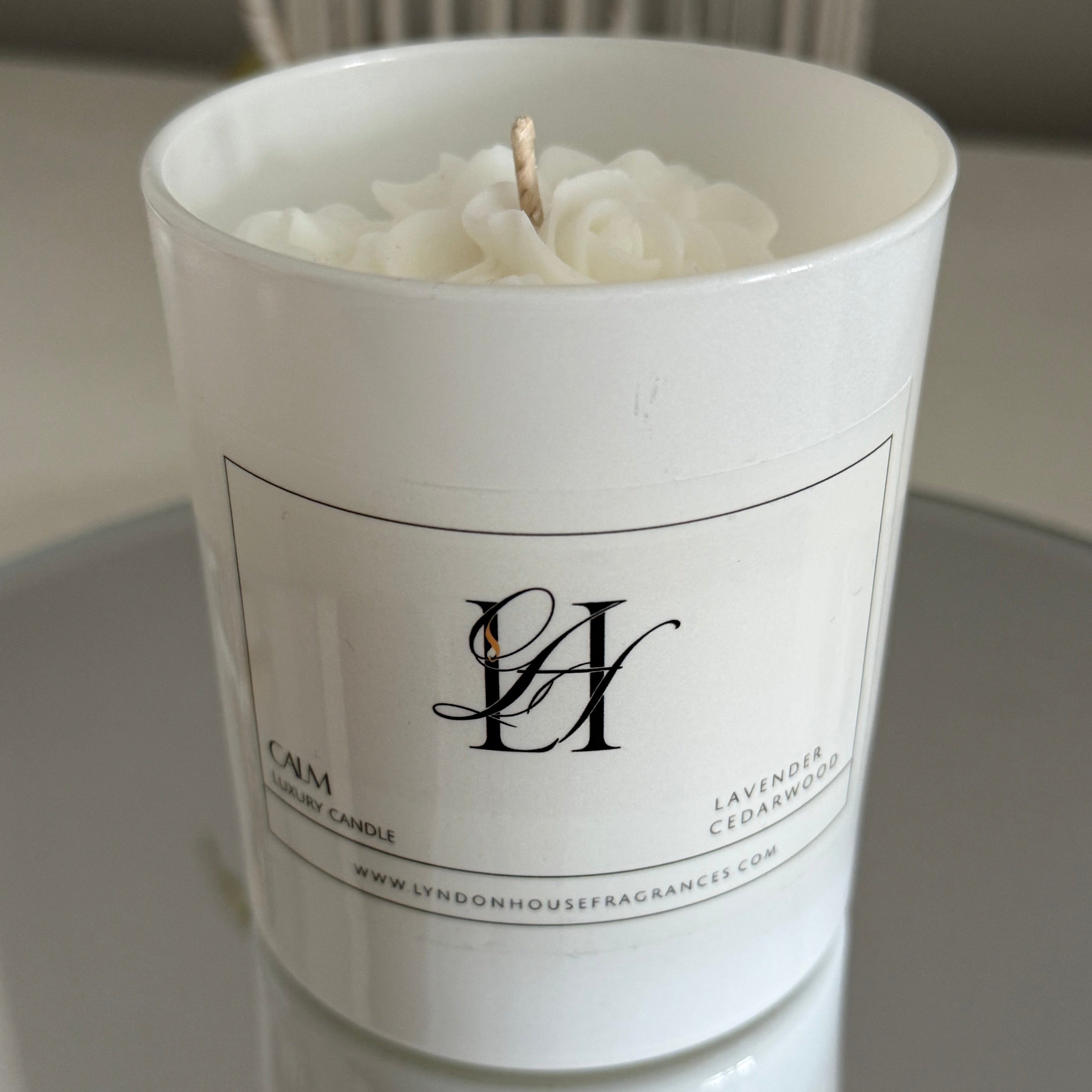 White candle in a cylindrical container with a brand logo on a neutral background
