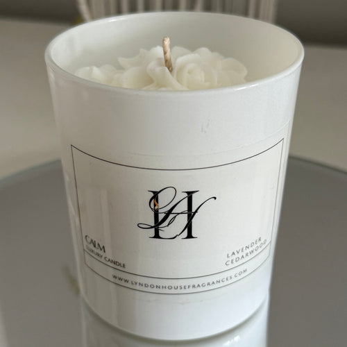 White candle in a cylindrical container with a brand logo on a neutral background