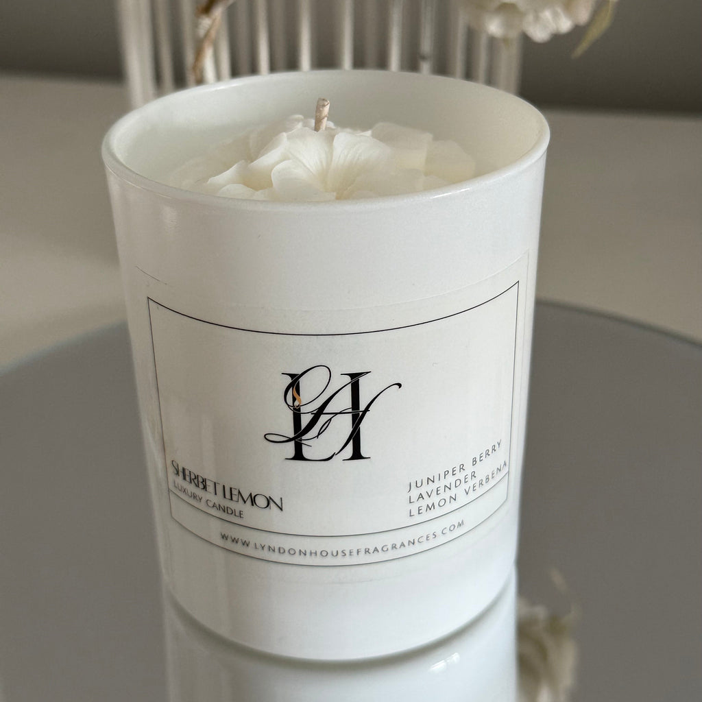 White candle in a cylindrical container with a label, placed on a surface with a blurred background.