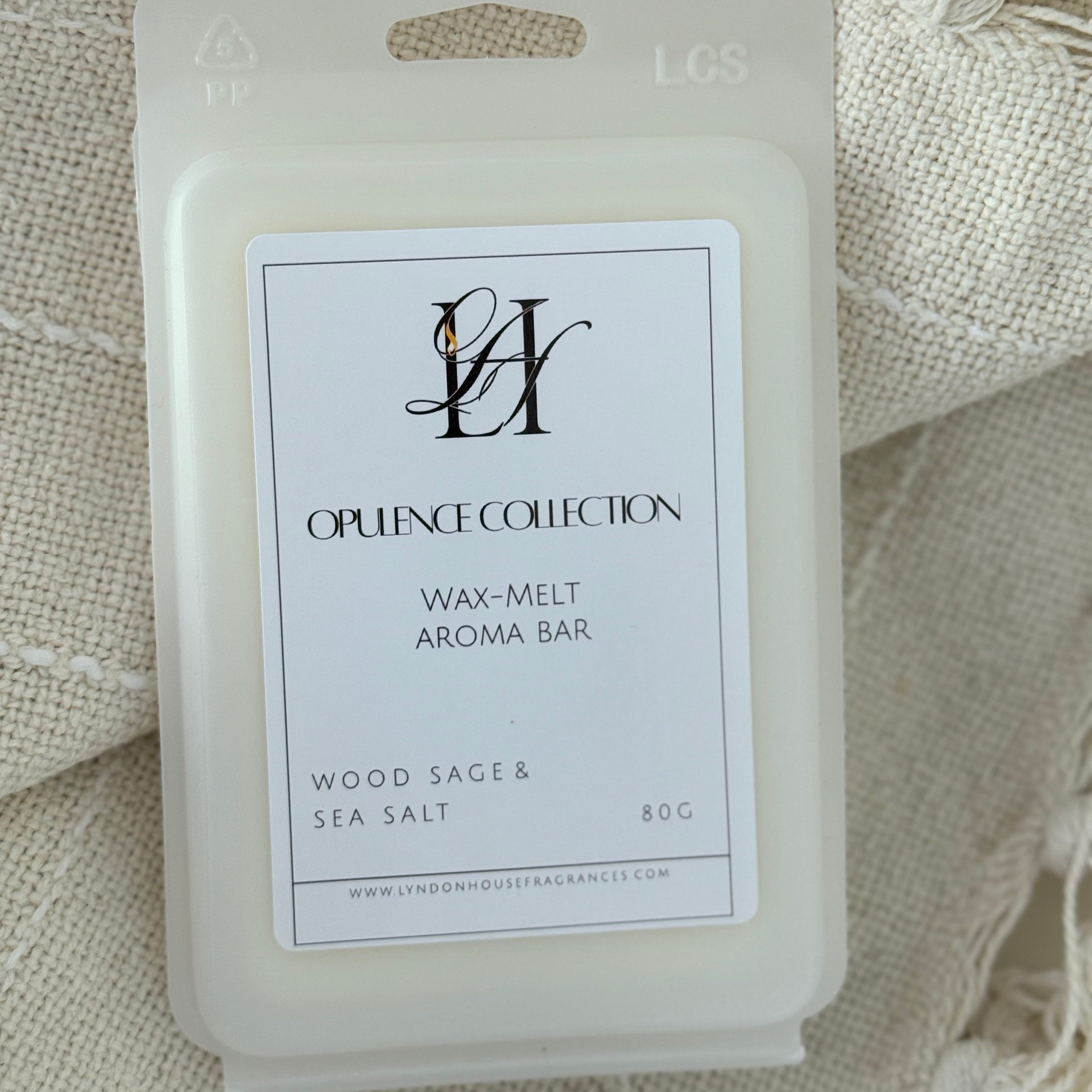 An 80g wax melt package from Lyndon House Fragrances Opulence Collection, featuring a cream-colored plastic clamshell with a white label that reads 'Wood Sage & Sea Salt'. The package is displayed on a textured, cream-colored woven blanket with tassels.