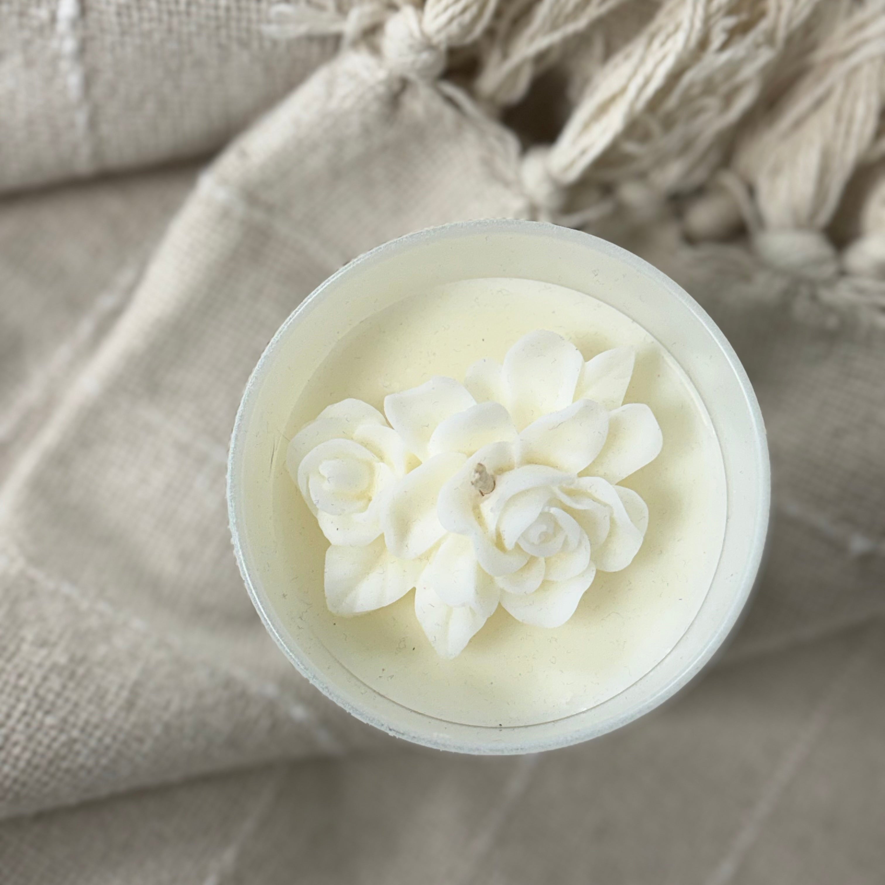 A top-down view of a white wax candle in a clear round container, decorated with a white flower design, resting on a beige textured fabric with tassels.